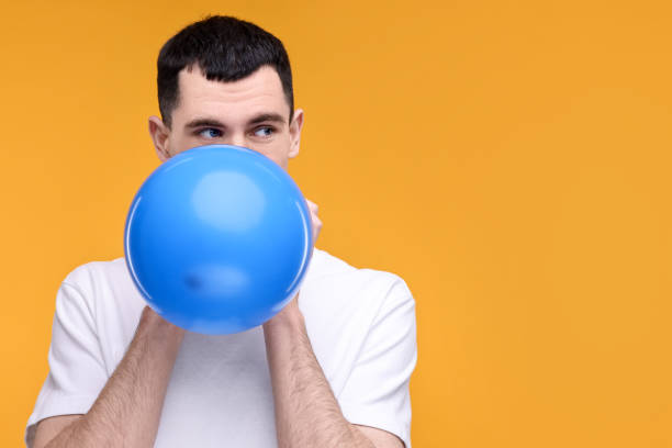 istock Young man inflating light blue balloon on yellow background. Space for text 2191428659
