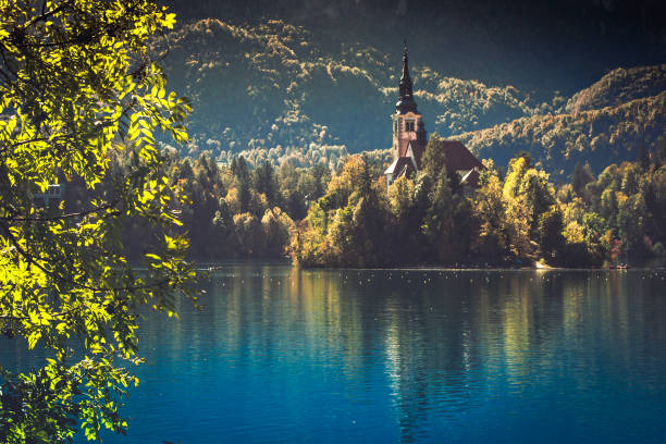 Lake Bled on a sunny autumn day. A view of the island and the Church of the Assumption of Mary. Traveling through Europe, Slovenia. A forest on the cliffs on a sunny day. Tourist attractions of Europe.
