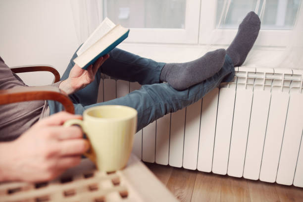 istock Man's feet with socks on a home heating radiator on a cold winter day outdoors while reading a book and drinking coffee tea from a cup. 2191445305
