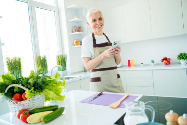 istock Elderly woman in kitchen apron smiling while preparing fresh meal using recipe on phone during sunny day indoors 2191449074