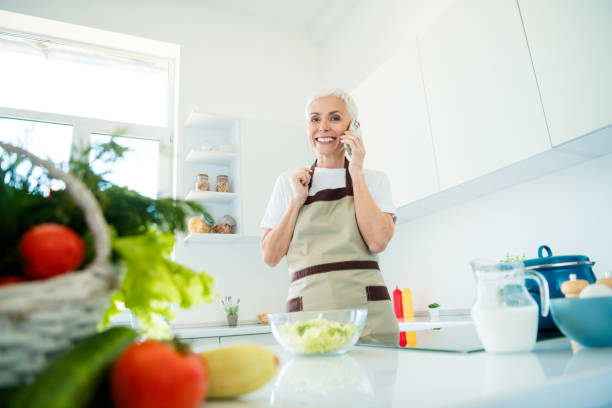 istock Charming mature woman in apron multitasking in bright kitchen, preparing meal while talking on phone, enjoying daylight and comfort 2191449142