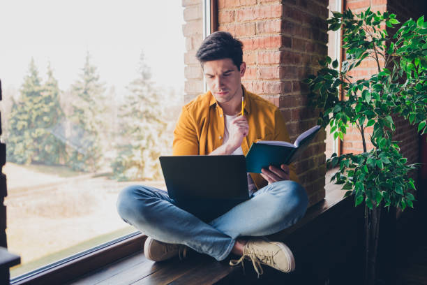 istock Young man relaxing at home, using laptop and reading a book by the window 2191449502