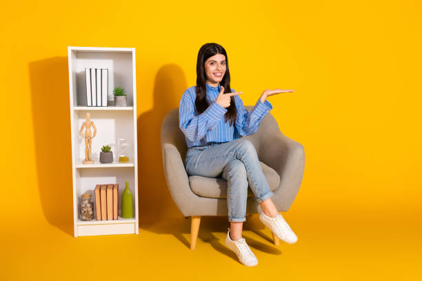 istock Young woman presenting something while sitting by a shelf with a yellow background 2191451107