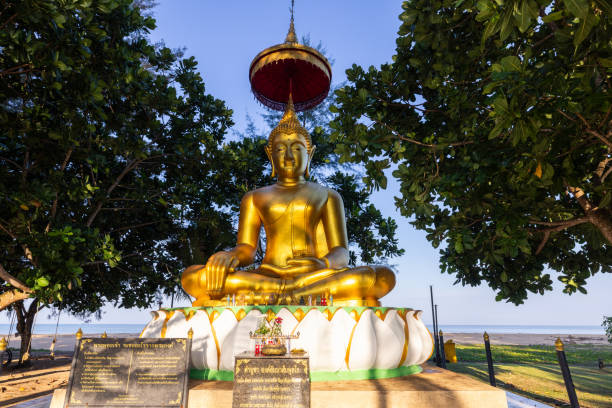 Golden Buddha statue sitting under lush trees in a serene outdoor setting. Peaceful religious shrine by the sea.