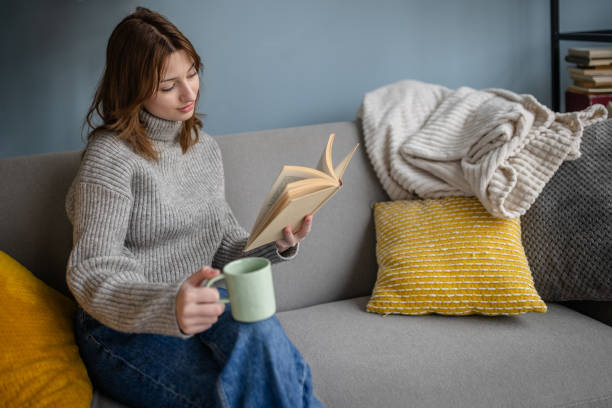 A young woman relaxes on a gray couch, with a book in one hand and a mug in the other, embodying the perfect cozy afternoon at home atmosphere.