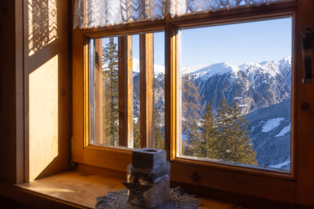 A warm cabin interior boasts stunning views of snow-capped mountains through large wooden windows on a crisp winter morning. The serene landscape invites tranquility.