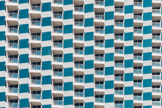 The image shows a close-up view of a building facade with a repeating geometric pattern. The building has balconies with glass railings, and the facade alternates between white and turquoise panels. The arrangement creates an optical effect, giving a sense of depth and movement.