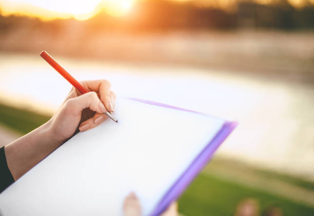 A close-up of a hand writing with a pen on a notebook during sunset, showcasing outdoor reflection and creativity, highlighting writing and documentation in a tranquil setting