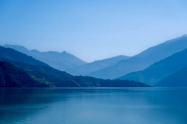 Multilayered blue mountains are shrouded in mist, with blue light cascading over them and the lake waters. This picturesque scene at Tehri Lake captures the enchanting interplay of light and nature.