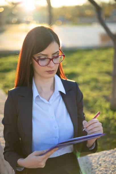 Young professional woman with red glasses writing on notepad in an urban setting, showcasing productivity and focus in a casual environment