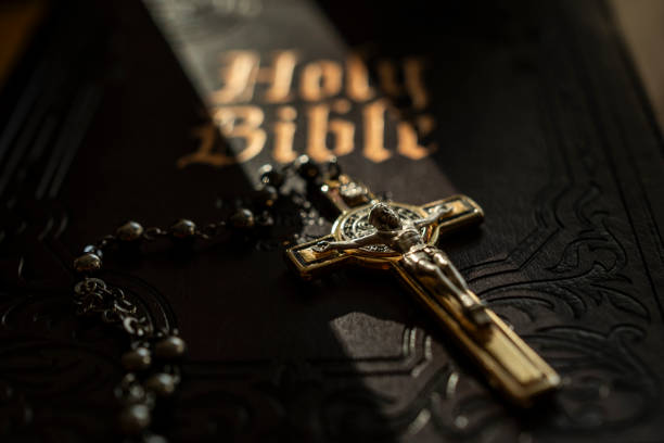 Close-up of a crucifix and rosary beads resting on an ornate Holy Bible cover, illuminated by dramatic light.