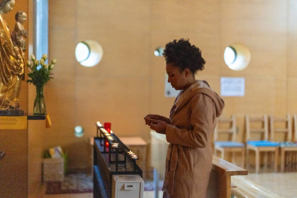 An African American woman standing in prayer inside an Adventist church, holding a small object in her hands, wearing a light-colored coat and a scarf