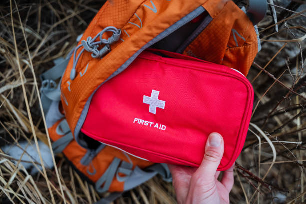 istock A person is holding a bright red first aid kit in front of a stylish orange backpack that they have placed on the ground, ensuring they are wellprepared for any emergency situations 2191489566