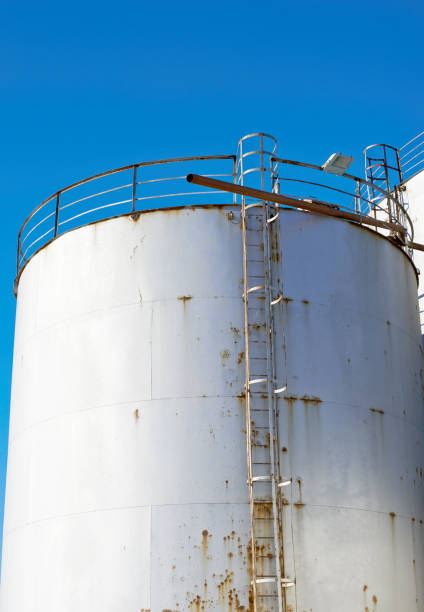 Prominent cylindrical white industrial storage tank with visible rust, metal ladder, and railing above, contrasting against a vibrant blue sky, symbolizing infrastructure, industry, and modern manufacturing.