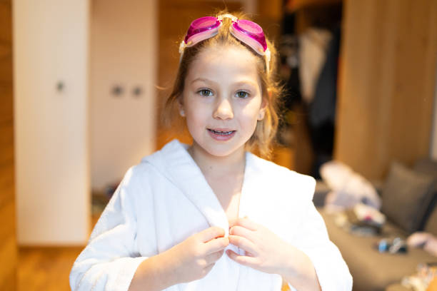 Portrait of cute little girl in bathrobe putting swimming goggles on while preparing for a swim in hotel room