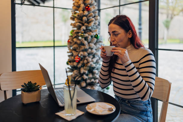 istock Female freelancer drinking coffee in a coffee shop and working remotely using a laptop. She is sitting by the window and next to the decorated Christmas tree 2191536016