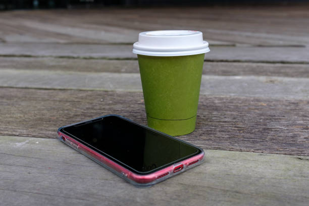 A take away green paper coffee cup with white plastic lid and a smart phone with black blank screen on wooden surface.