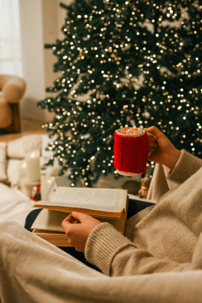 istock A cozy holiday scene featuring a person reading an open book while holding a festive red mug filled with marshmallows 2191545113
