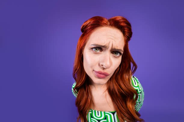 A fashionable young redhaired woman showing a curious expression, standing against a bright violet background. The image emphasizes contemporary style, charm, and a playful attitude with a trendy outfit.