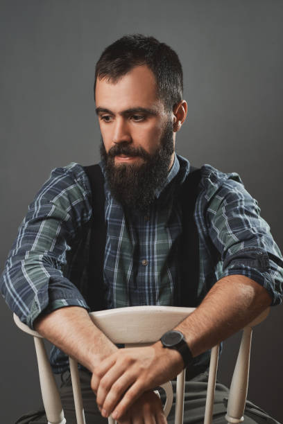 A young and handsome man poses on a chair in a studio in front of a dark gray wall.
