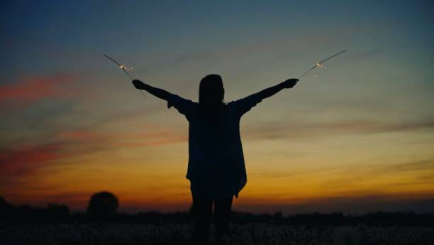 Caucasian woman in casual attire celebrates with sparklers on agricultural field during sunset, creating dramatic silhouette against golden sky.