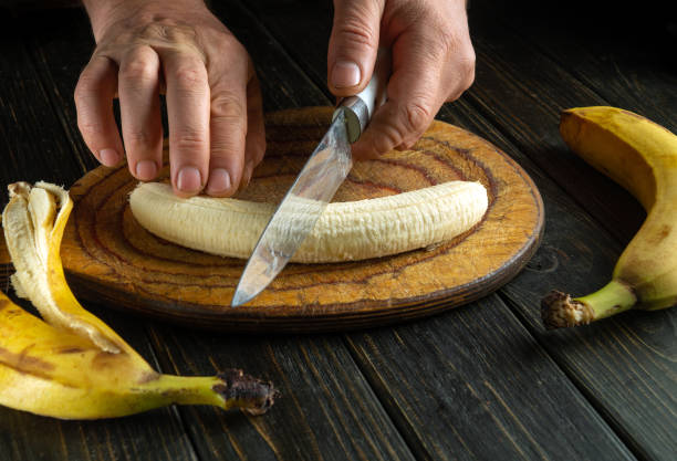 istock Hands skillfully cut a fresh banana on a rustic wooden board, surrounded by ripe bananas. The warm atmosphere invites a sense of home and culinary creativity 2191562607