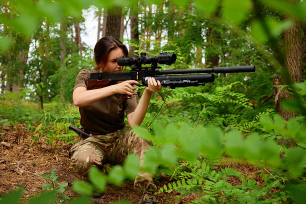 istock A marksman is positioned low among ferns and trees, focusing intently through a scope as they prepare to shoot. The environment is rich with greenery and natural light 2191563313