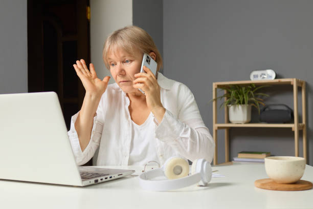 istock A middle-aged woman sits at a desk, talking on the phone with one hand while using her laptop. Her home office is well-lit and decorated with plants and books 2191563368