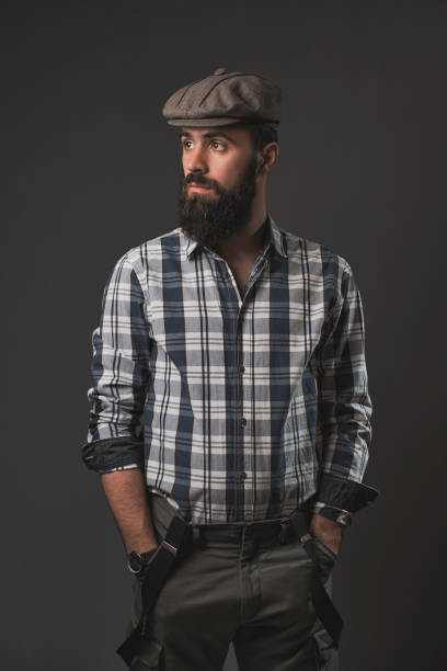 Young handsome man posing in the studio in front of a gray wall. Cap, plaid shirt, beard, hipster style.