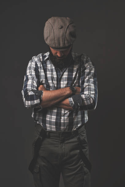 Young handsome man posing in the studio in front of a gray wall. Cap, plaid shirt, beard, hipster style.