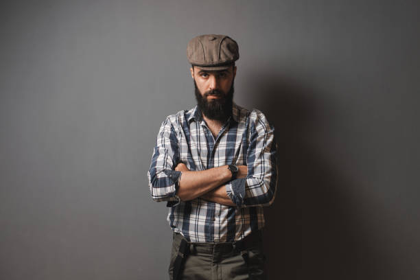 Young handsome man posing in the studio in front of a gray wall. Cap, plaid shirt, beard, hipster style.