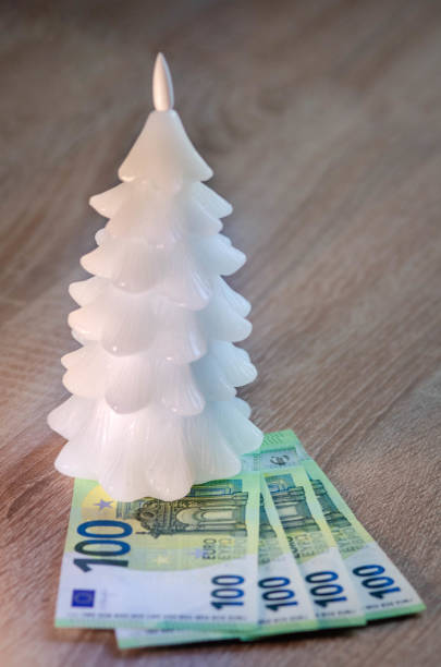 A small white Christmas tree ornament with stacked 100 euro banknotes represents the expenses we have during the New Year -stock photo Small candle ornament and stacked hundred euro banknotes in cash represents the expenses we have during the New Year and Christmas holidays for gifts and decorating the space. euro bill in a christmas ornament stock pictures, royalty-free photos & images