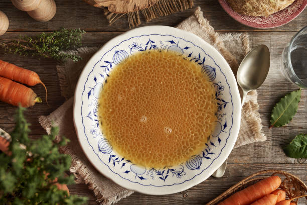 Chicken bone broth in a white plate with blue ornaments with fresh vegetables, top view