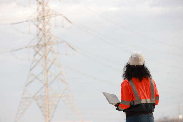 A Black female electrical engineer using a laptop computer to stand at the electricity station to see the electric power production plan at a high voltage electrode at the pier