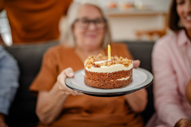 Close-up view of a cake held by an elderly woman in a living room, featuring a lit candle as the focal point, capturing a moment of joy and celebration.