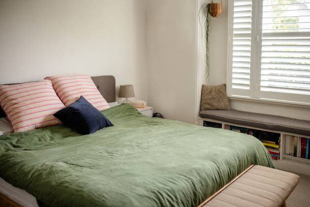 A inviting bedroom featuring a plush green duvet, several pillows in different shades, soft window lighting, and a bench near the window seat for reading.