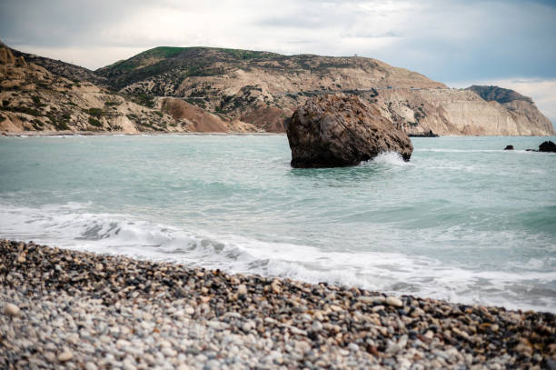 A scenic coastal landscape featuring a rocky beach with a large boulder in the sea, surrounded by turquoise waters and distant hills under a cloudy sky.