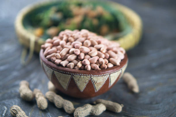 A bowl filled with shelled peanuts displayed on a textured wooden table with shells and blurred greens in the background, perfect for themes of nutrition, rustic style, or culinary ingredients