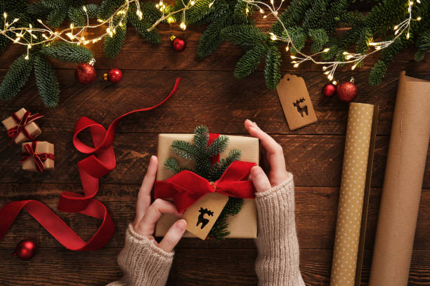 istock Preparing for the holiday, gift wrapping. Women's hands attach a tag to a gift box. Table with handmade items, rolls of kraft brown paper, red velvet ribbon, labels, scissors. 2191595615