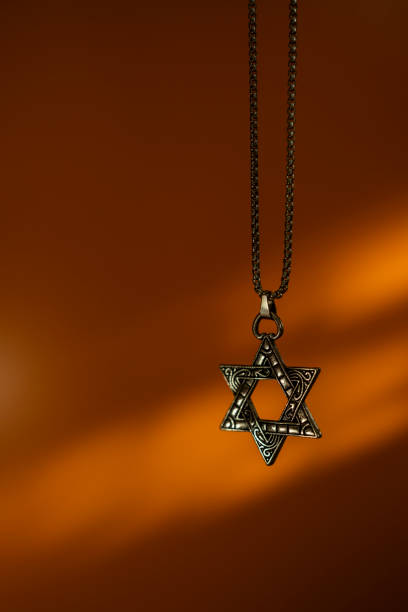 Close-up of a metallic Star of David necklace, set against a warm orange background with soft lighting.