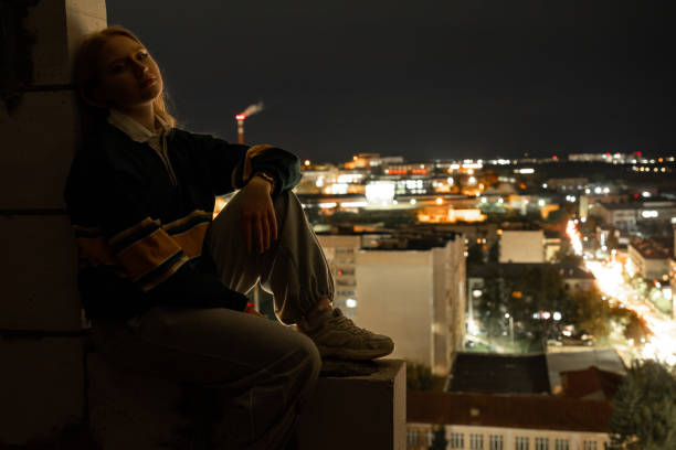 Young woman sitting on a rooftop at night, gazing at illuminated city lights below.