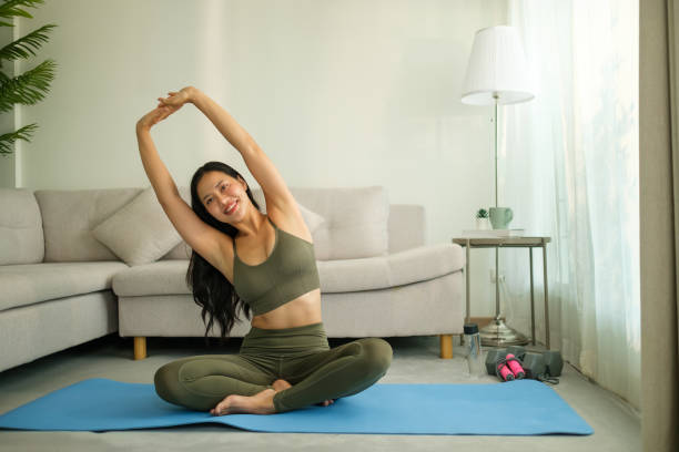 istock Smiling woman practicing yoga at home, seated on a blue mat in a cross-legged position while stretching her arms overhead. 2191620368