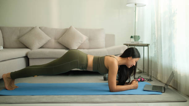 istock Woman in activewear performs a plank exercise on a yoga mat while watching a fitness tutorial on her laptop in a cozy living room setting. 2191620402