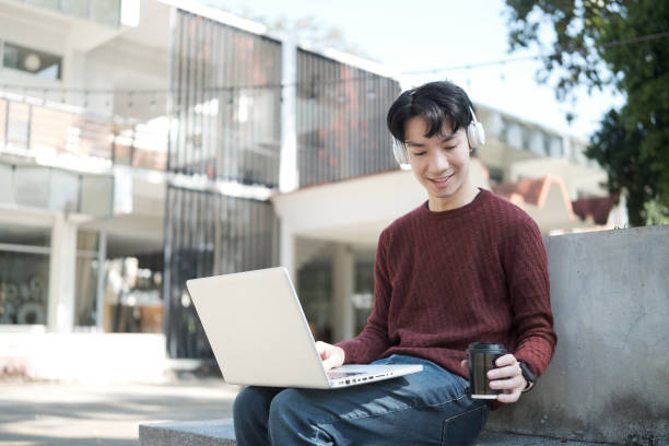 istock A young man sitting outdoors with a laptop on his lap, wearing a maroon sweater and white headphones. 2191620499