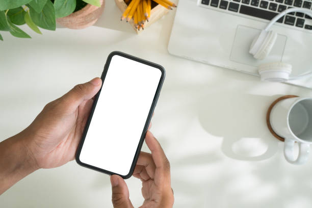 istock Close up of hands holding a smartphone with a blank screen, for showcasing app designs or user interface concepts, set on a minimalist desk with a laptop, headphones and coffee mug. 2191620643
