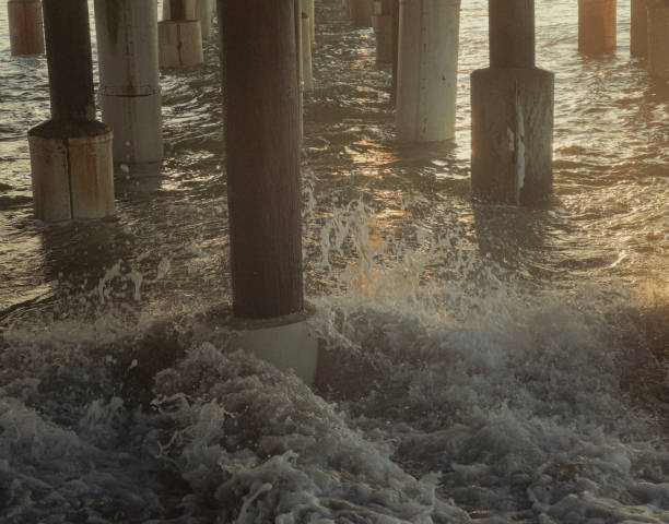 Dramatic view of the ocean waves crashing against the underside of a weathered wooden pier structure. The sunlight filters through the gaps in the pier, casting a dappled light effect on the water below. American flag hangs off the pier.