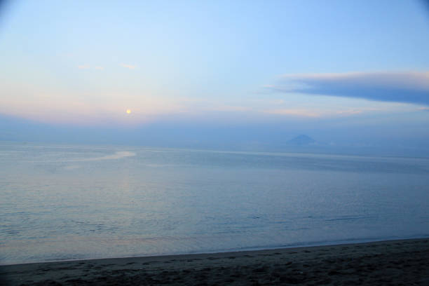 A tranquil beach scene during twilight, with the moon casting a soft reflection on the calm ocean. The horizon features a distant mountain peak partially obscured by clouds, under a pastel-toned sky.