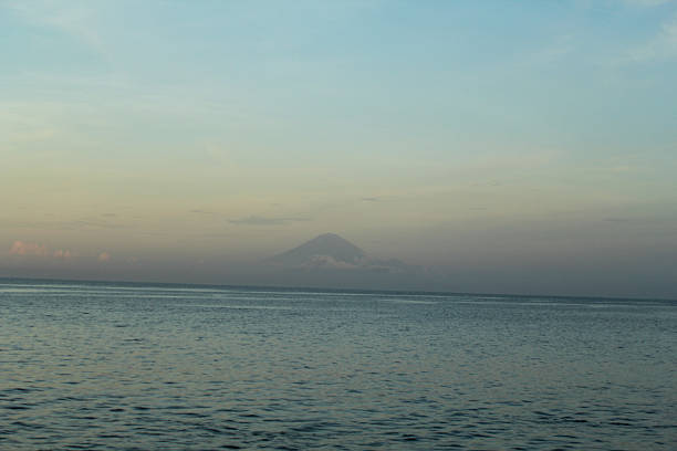 A tranquil view of the ocean at dusk with a distant volcano, partially veiled by clouds, silhouetted against a pastel-colored sky blending into soft blue waters.