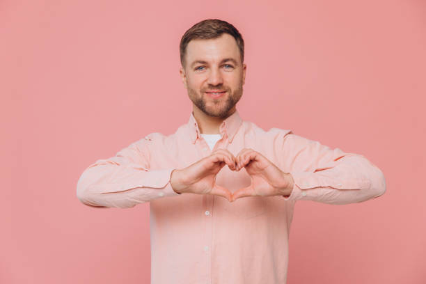istock Lovely good-looking caucasian bearded guy smiling cheerfully and making heart gesture over chest, expressing his feelings and tenderness while standing against pink background. I am in love with you 2191658366