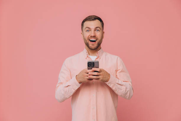 istock Handsome attractive smiling guy millennial hold telephone hands popular blogger addicted user wear casual pink shirt isolated pink color 2191658424